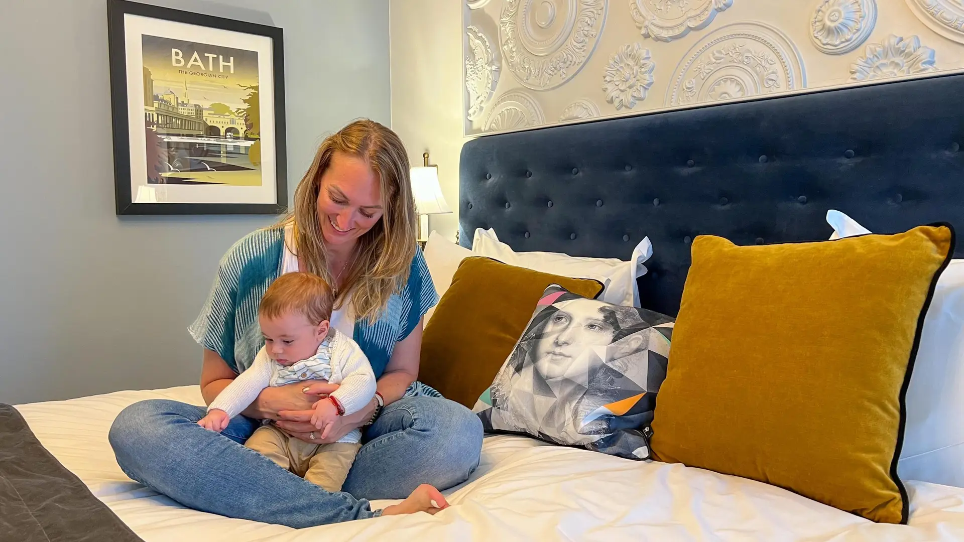 A woman sits on a bed, holding and smiling at a baby. The bed has green and beige pillows, and a framed picture of Bath on the wall behind them. This family-friendly accommodation offers affordable comfort and charming decor perfect for making lasting memories.