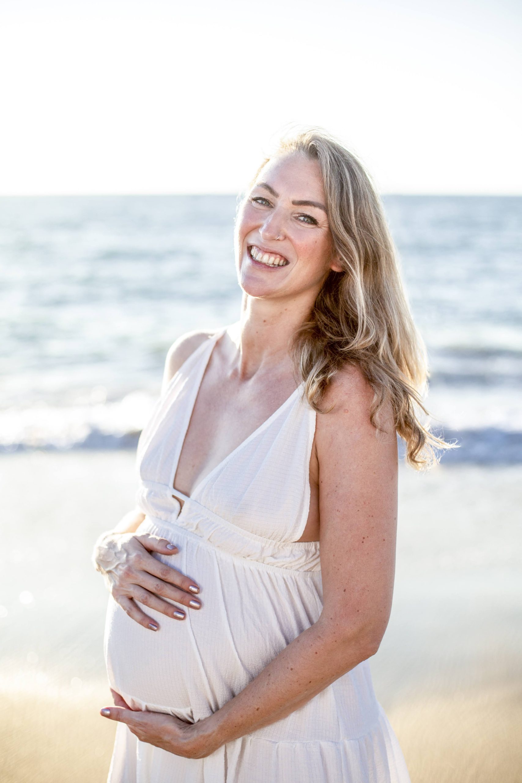 A smiling pregnant woman in a white dress stands on a sunny beach, holding her belly with both hands.
