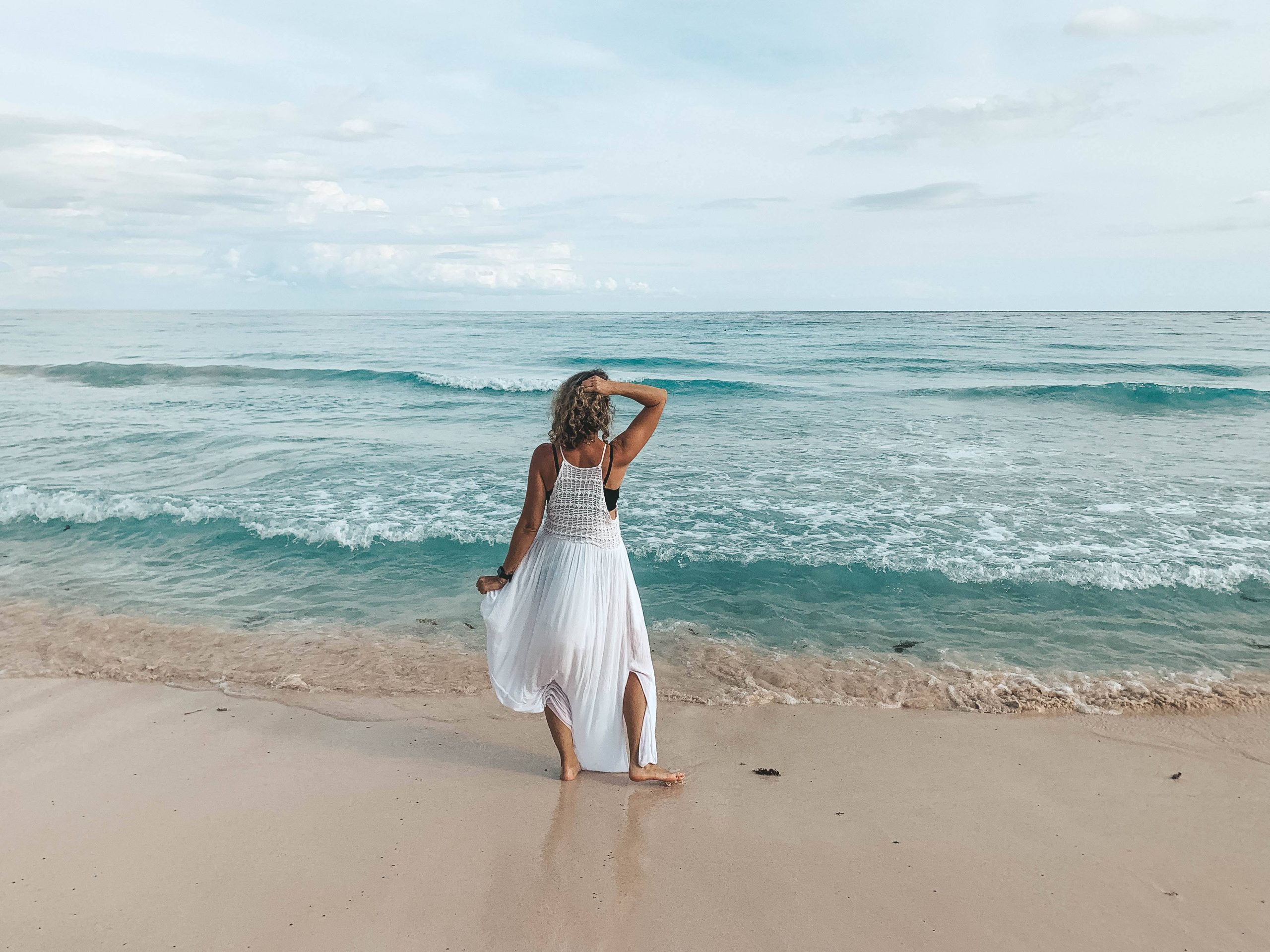 A person in a white dress stands on a sandy beach facing the ocean, with one hand on their head and waves gently crashing in the background.