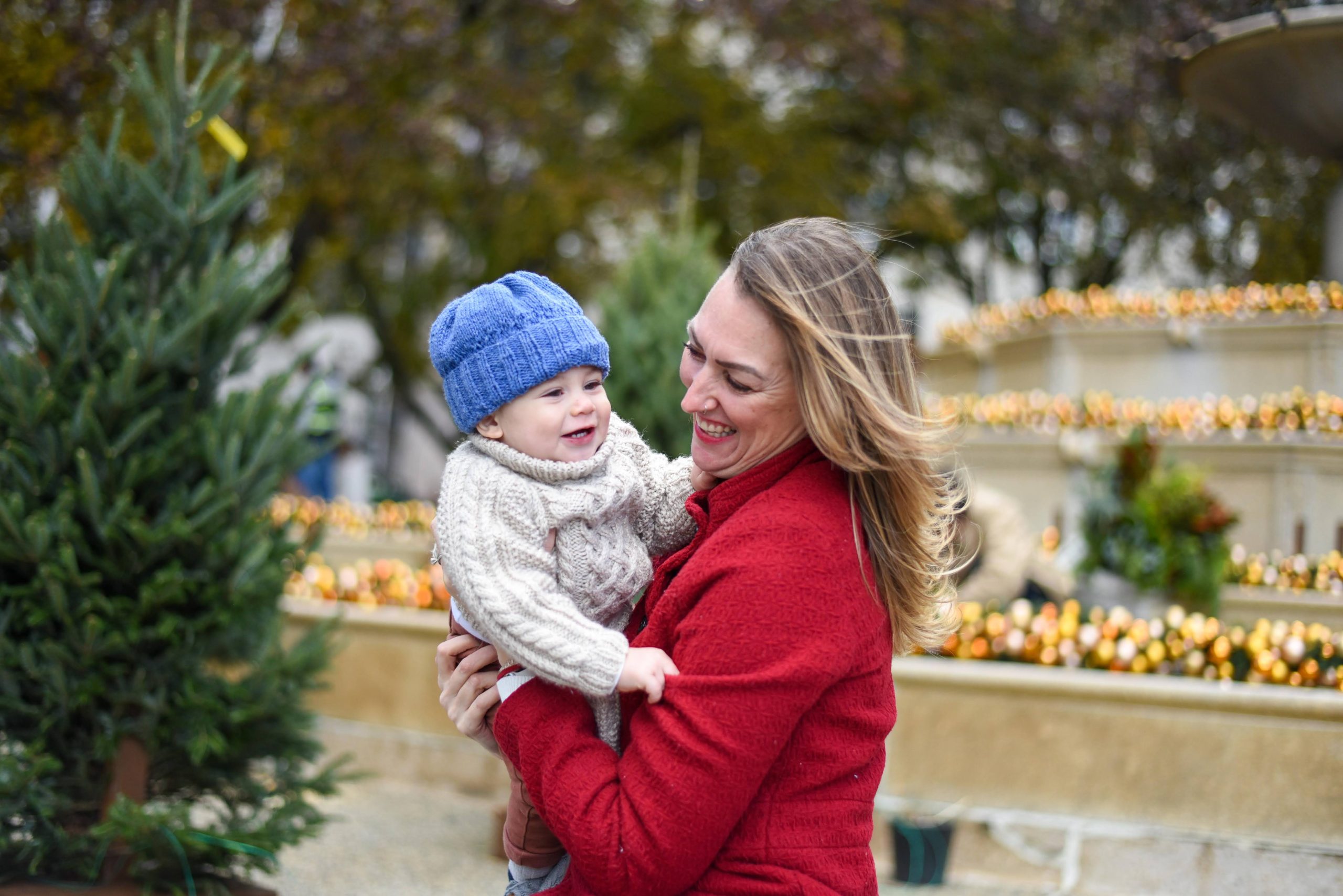 A woman in a red coat smiles while holding a child wearing a blue hat and beige sweater. They are outdoors with decorative lights and pine trees in the background.
