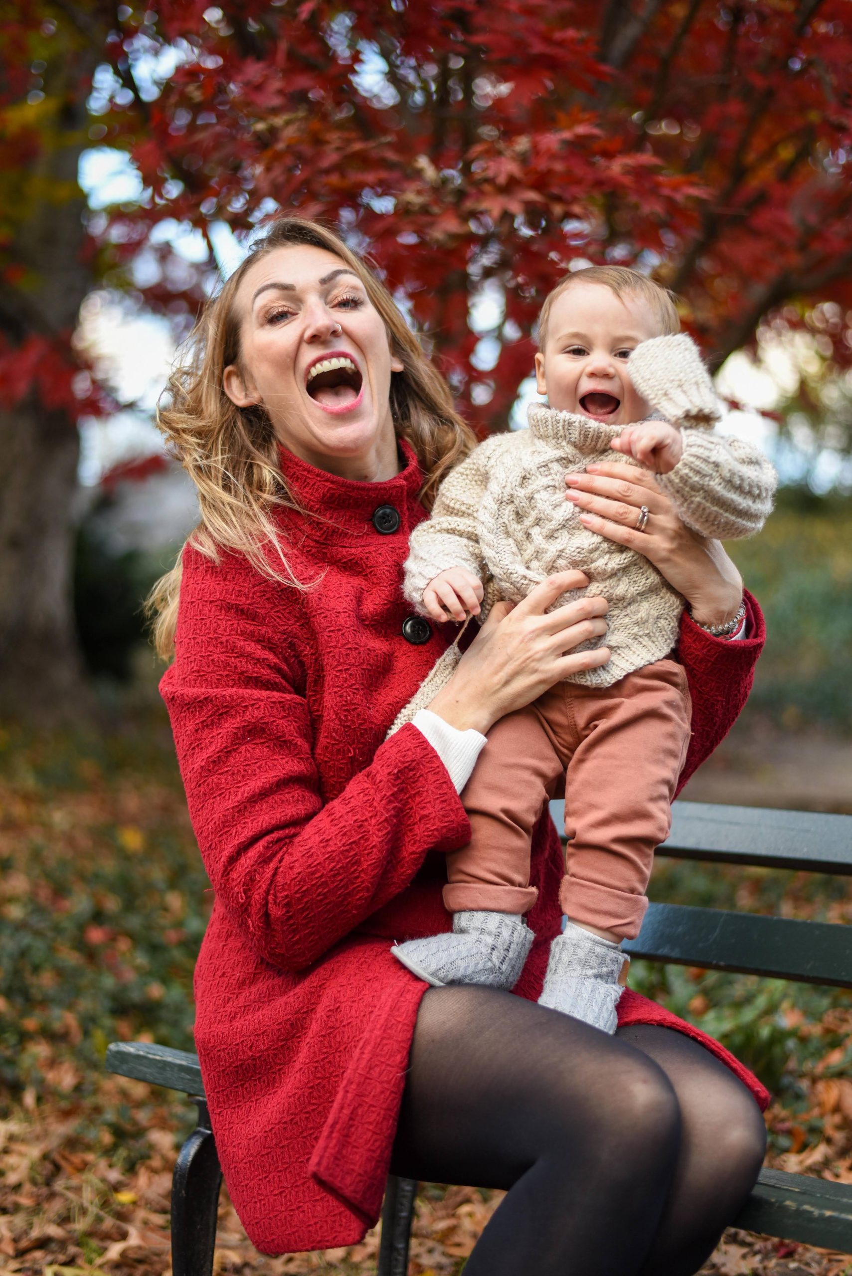 A woman in a red coat sits on a bench outdoors, holding a baby in brown pants and a beige sweater. They are both smiling and surrounded by autumn foliage.