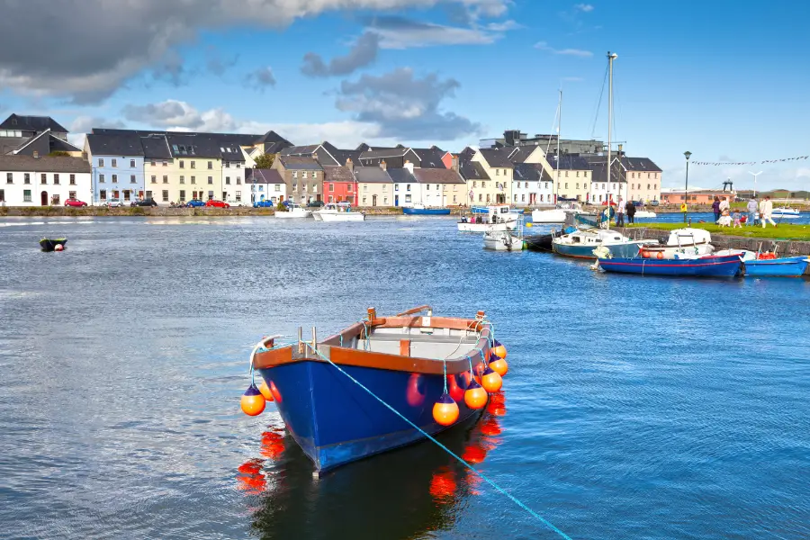 A blue boat with orange buoys floats in a Galway harbor, with several other boats docked nearby. Colorful houses line the waterfront under a partly cloudy sky, making it an ideal spot for family things to do.