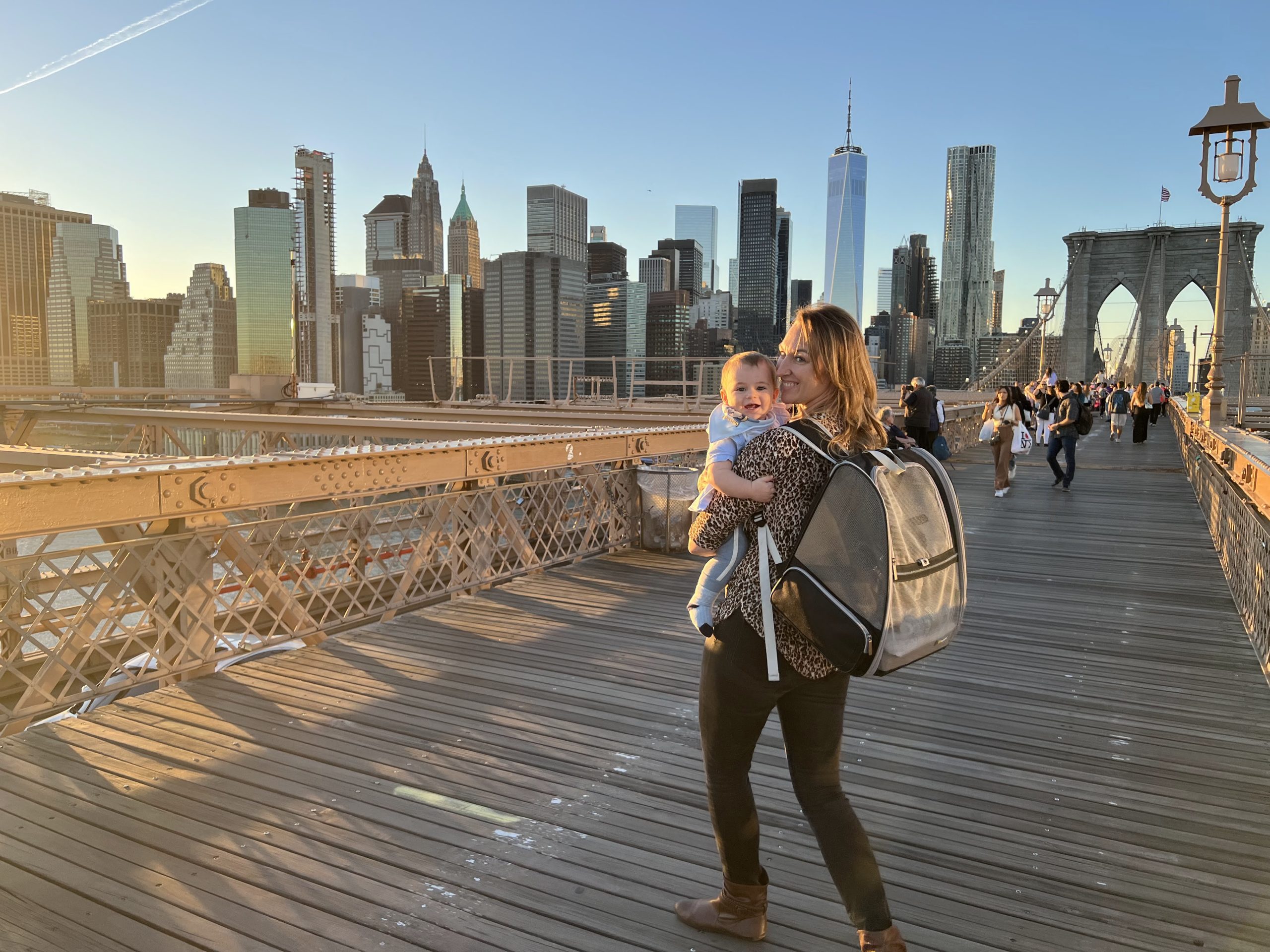 A person holds a baby while standing on the Brooklyn Bridge with the New York City skyline in the background during sunset.