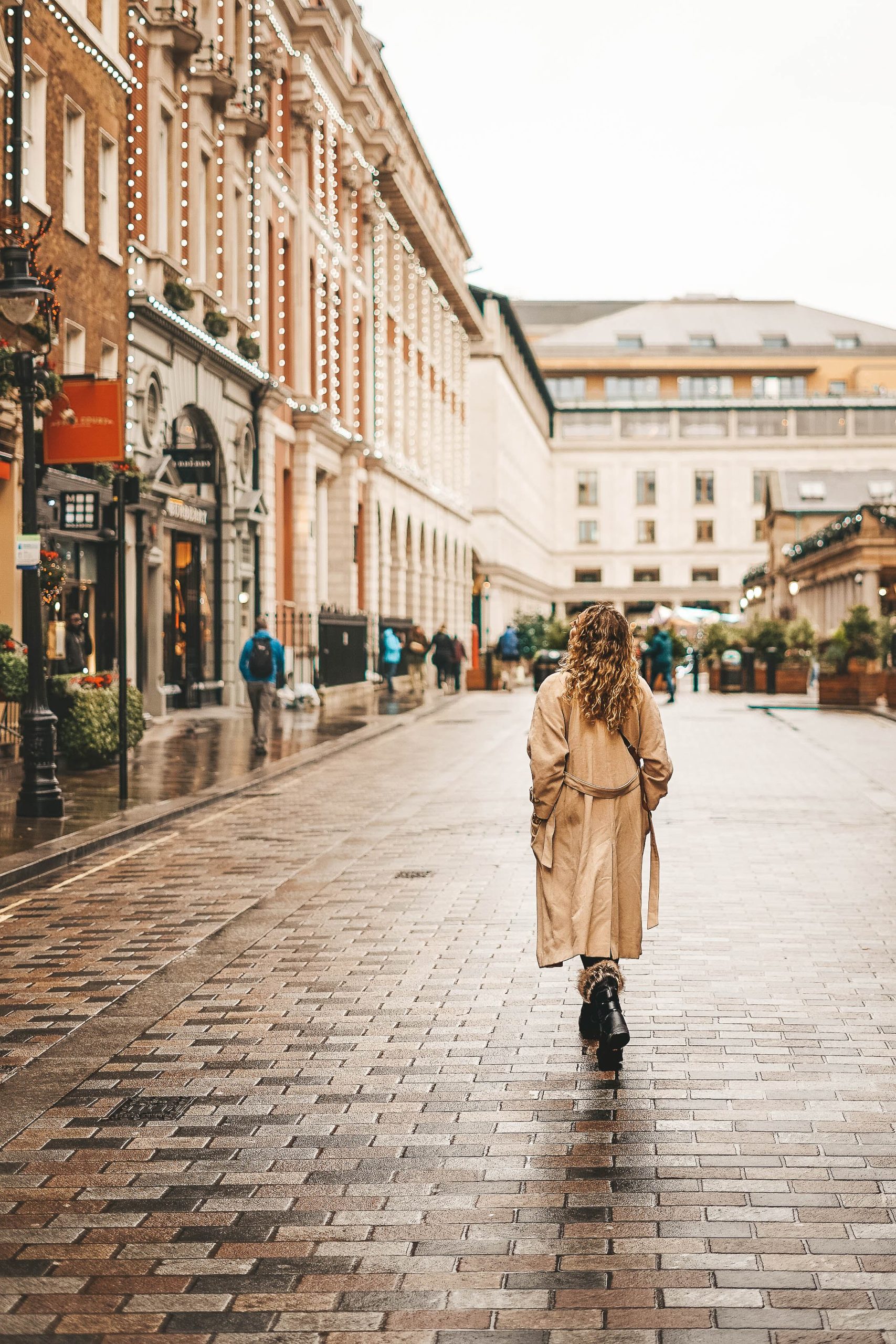 A person with long curly hair wearing a beige trench coat walks down a cobblestone street lined with buildings and shops.