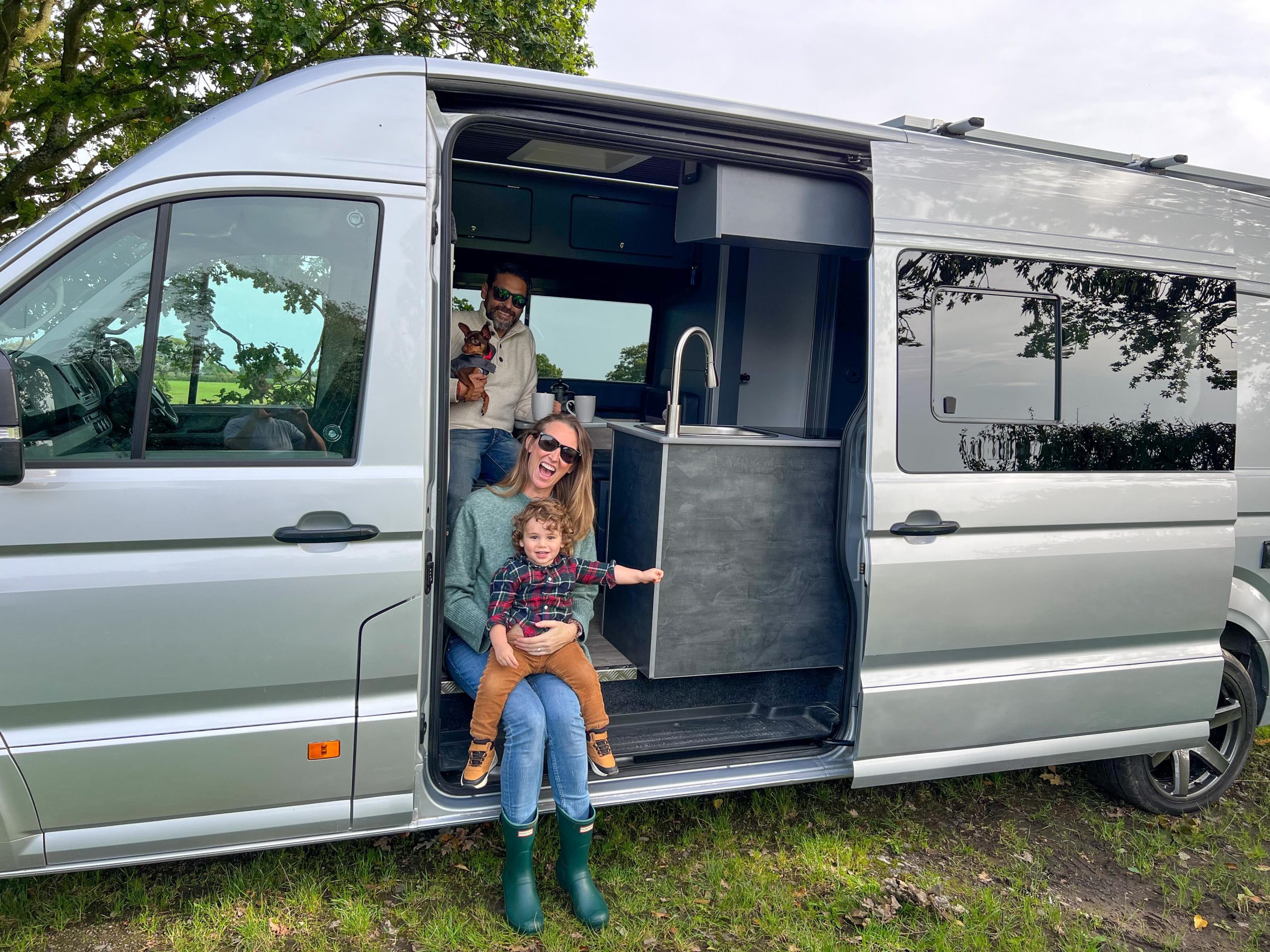 A family sits in and around a silver camper van parked on grass, with a man holding a small dog inside, a woman sitting on the step, and a child standing next to her.