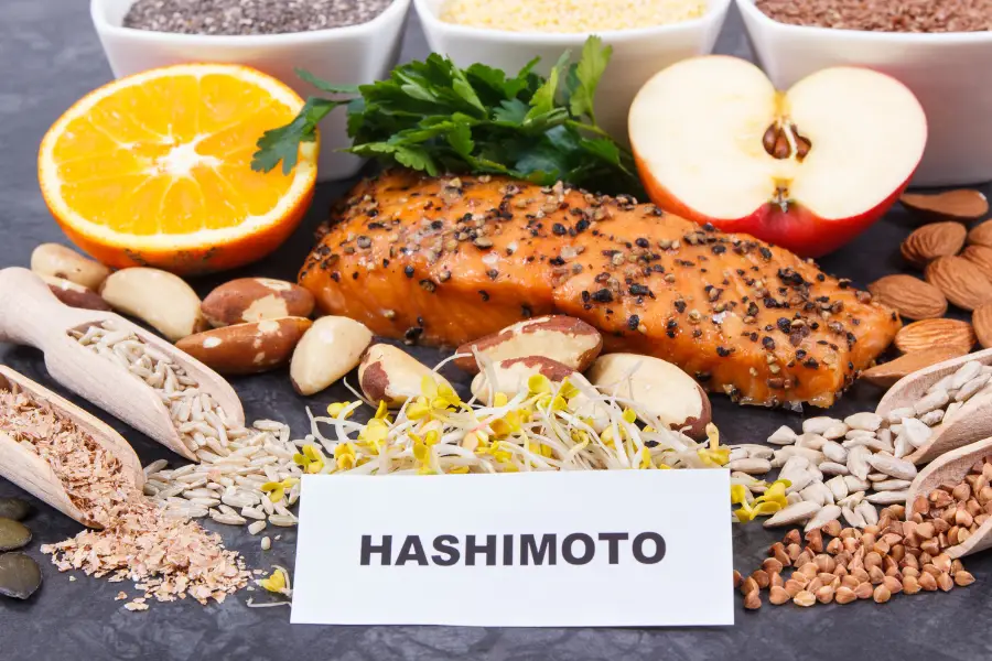 A variety of foods, including salmon, chia seeds, nuts, an orange slice, an apple half, sprouts, and leafy greens, are arranged on a surface with a card labeled "Hashimoto." This visually appealing spread could easily feature in an Ayurveda beginners guide.