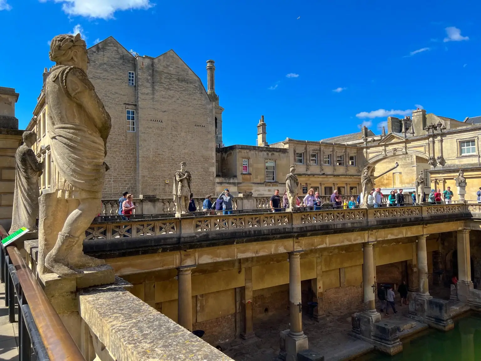 A historic building with statues and people walking on a stone walkway, featuring a pool below, under a bright blue sky—perfect for family-friendly activities.
