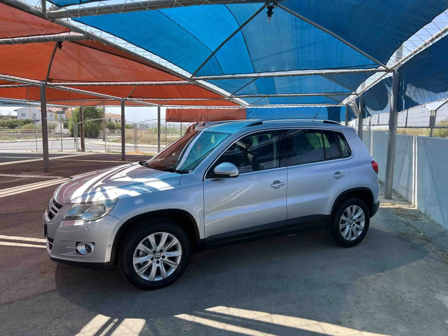 A silver SUV is parked under a canopy with a combination of blue and orange shades. The ground is paved, and the background shows a fence and a distant building. If you're considering how to buy a car, this scene might inspire your vision for buying a car in Cyprus.