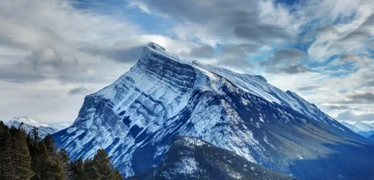 Snow-covered mountain under a cloudy sky with patches of blue. The landscape includes some pine trees in the foreground, perfect for a family-friendly ski vacation.