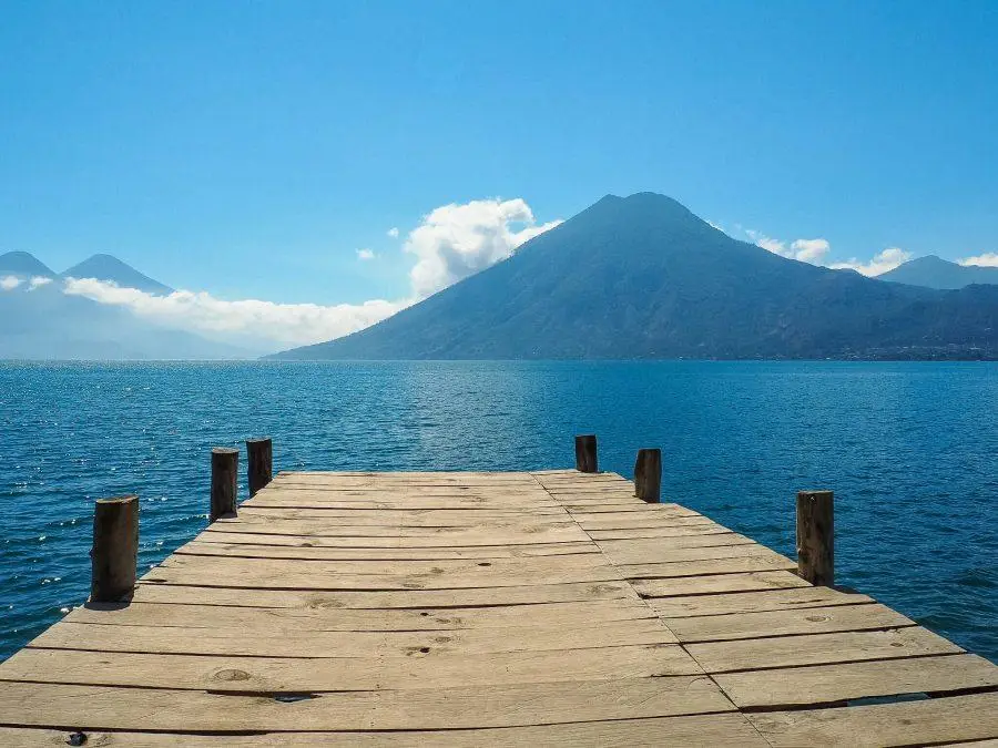 A wooden dock extends into a calm lake with mountains visible in the background under a clear blue sky, reminiscent of a serene evening in Guatemala.