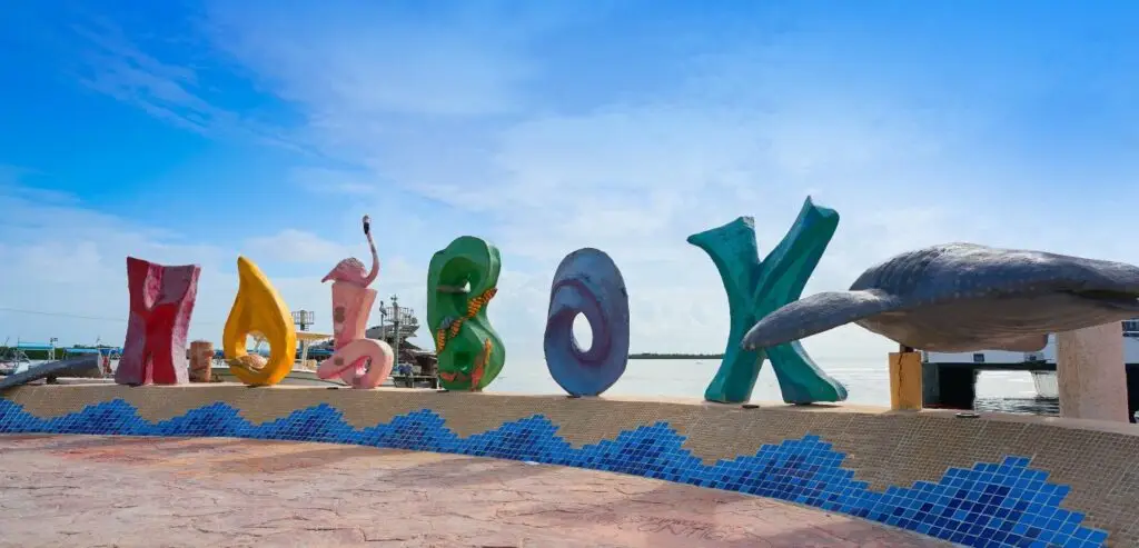 Colorful large letters spelling "Holbox" stand along a waterfront promenade with a whale shark sculpture on the right side. The background features a cloudy sky and calm water, making it one of the best things to do in Isla Holbox with kids for memorable family activities.