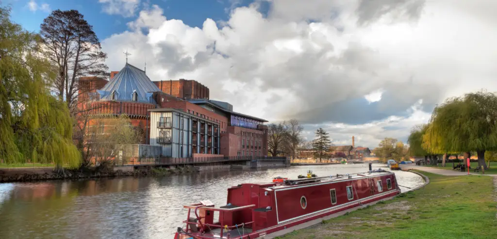 A red narrowboat is moored on a canal next to a modern, brick building with a unique roof design in Stratford Upon Avon. The area is surrounded by lush greenery and a cloudy sky, offering plenty of things to do for children and families exploring the scenic views.