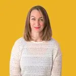 A woman with shoulder-length brown hair and red lipstick wears a white knit sweater while standing against a solid yellow background, looking like she's ready to grace the home page of a fashion magazine.