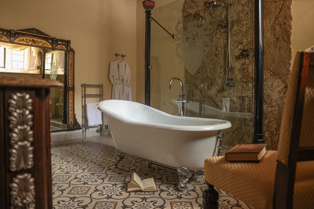 A vintage-style bathroom reminiscent of a Tudor Castle, featuring a freestanding bathtub, decorative tile flooring, a glass shower, and an ornate mirror. A robe hangs beside a chair with books and a towel on it, evoking the charm of Thornbury Castle.