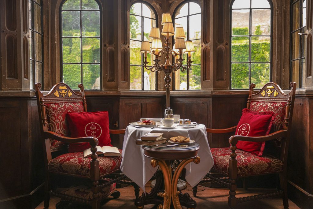 Two ornate chairs with red cushions flank a small round table set with a white tablecloth, tea, and snacks. A tall candelabra lamp stands in front of large windows overlooking the greenery of Thornbury Castle, evoking the charm of a night in a Tudor castle.
