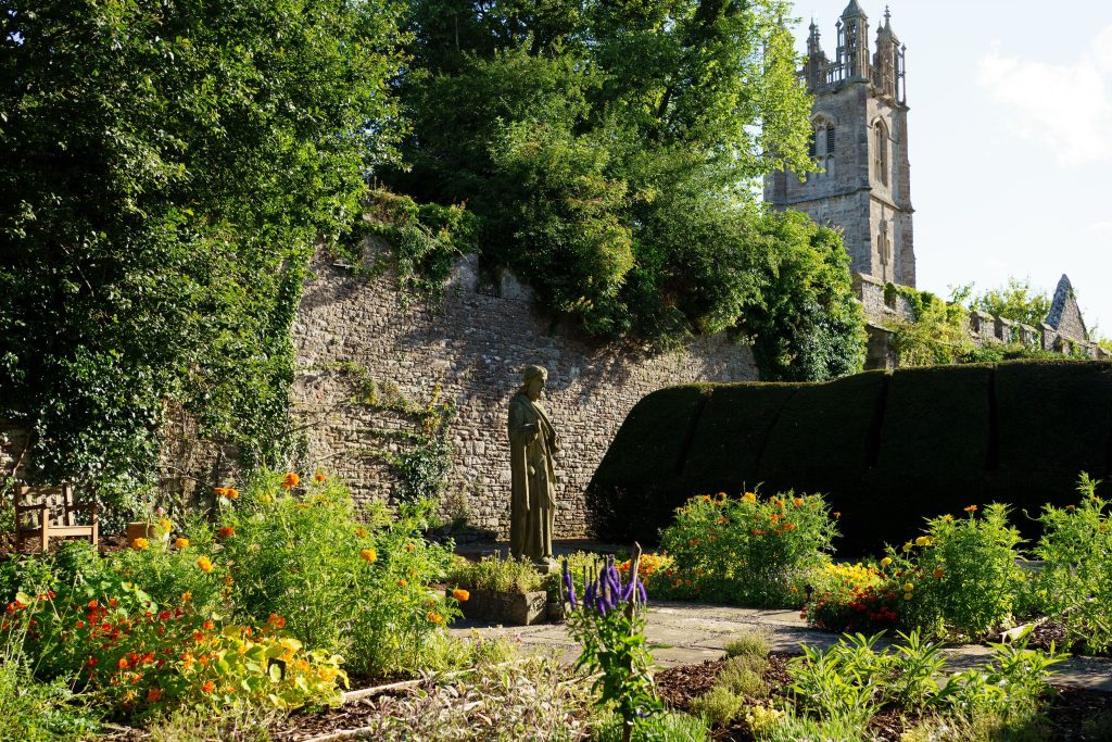 A statue of a cloaked figure stands in a garden with colorful flowers, reminiscent of the enchanting grounds at Thornbury Castle. A stone wall and a tall church tower rise elegantly in the background, completing this picturesque scene.