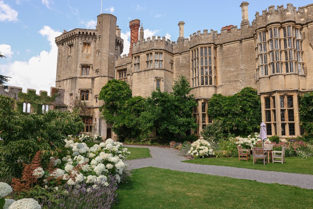 Historic Tudor castle with crenelated towers, tall windows, and ivy-covered walls. Manicured garden with blooming white flowers, pathway, and wooden chairs in the foreground under a blue sky at Thornbury Castle.