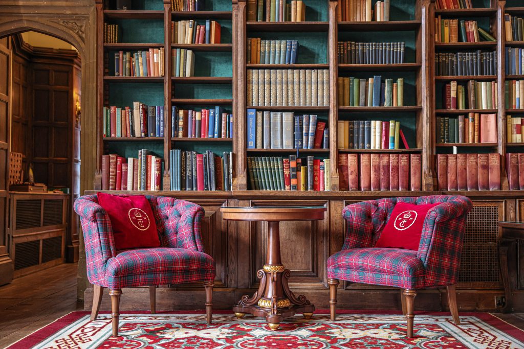 Two plaid armchairs face a round wooden table, set against a large bookshelf filled with books in multiple colors. A patterned rug covers the floor, evoking the cozy charm of a night stay at Thornbury Castle.