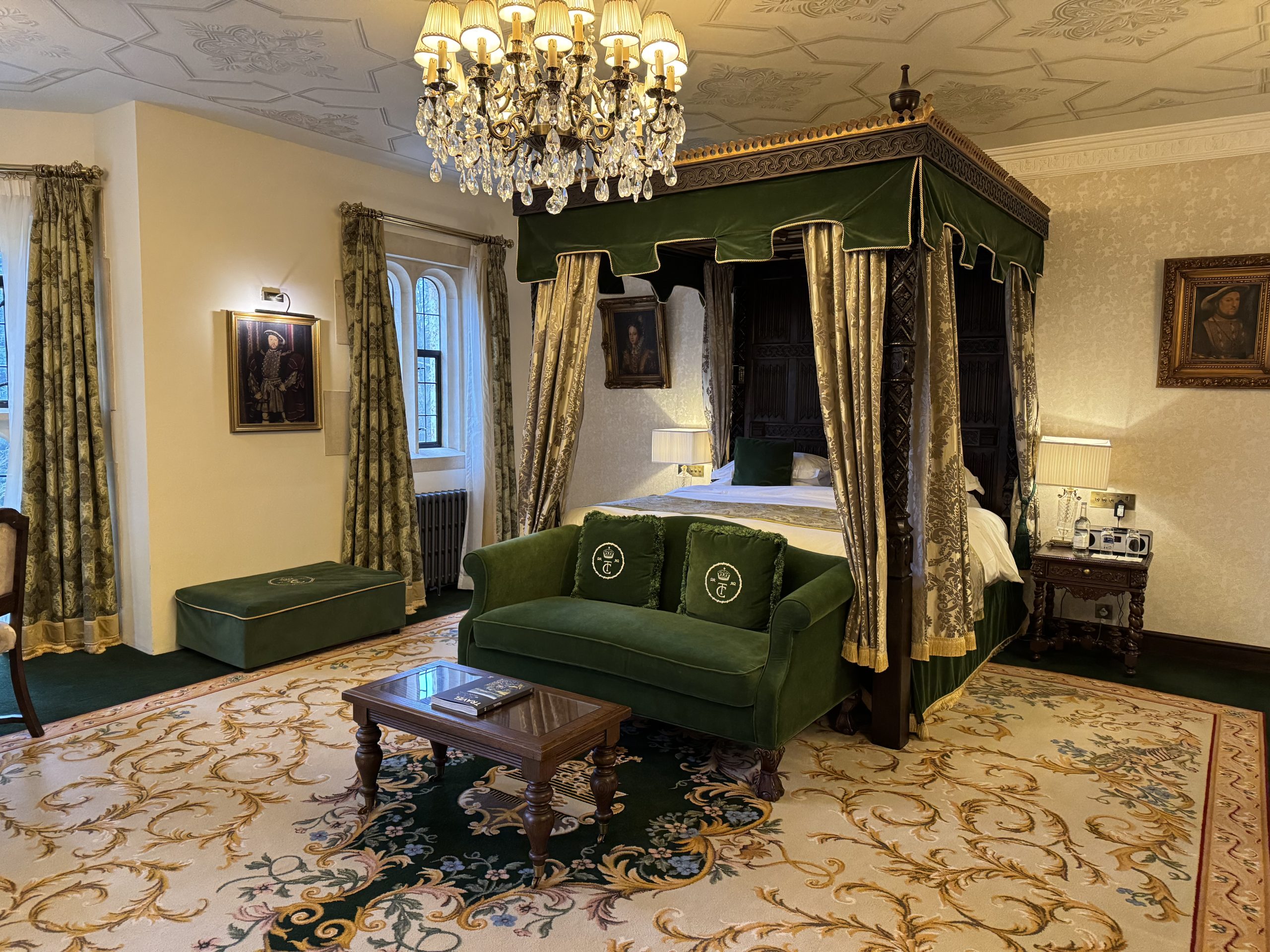 Luxurious room reminiscent of a Tudor Castle, featuring a green canopy bed, matching sofa, and ornate carpet. A crystal chandelier hangs from the ceiling while two framed paintings adorn the walls, evoking the elegance of Thornbury Castle.