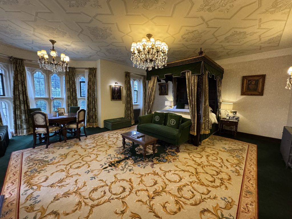 Experience a night in a Tudor Castle with this luxurious room featuring a canopy bed, green and beige decor, elegant chandeliers, ornate carpet, and arched windows adorned with green drapes. Feel the grandeur reminiscent of Thornbury Castle.