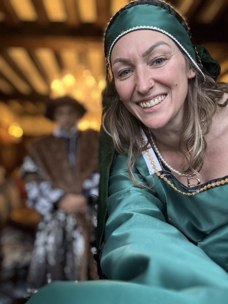 A woman in a green medieval costume smiles at the camera, capturing the enchanting allure of Thornbury Castle. In the background, a figure in historical attire adds to the Tudor charm of the scene.