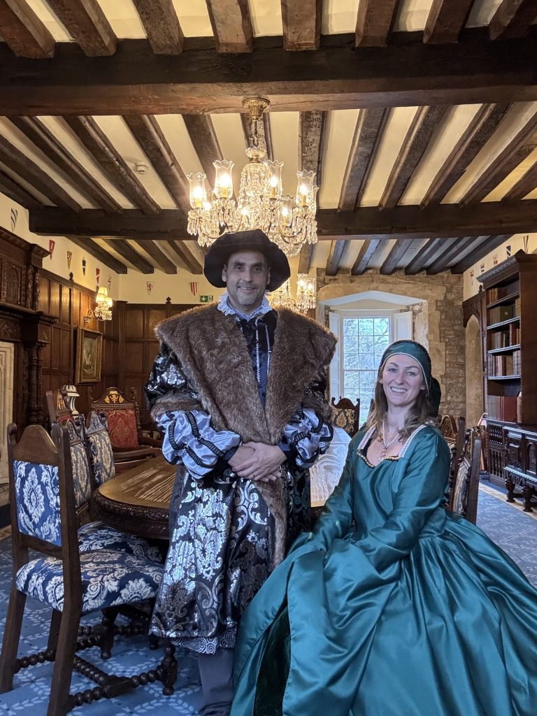 Two people in historical costumes sit in a richly decorated Tudor Castle room, complete with wooden beams, chandeliers, and a large window as the backdrop.