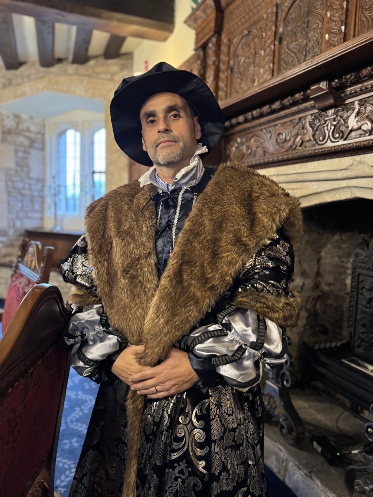 In a room with stone walls and a cozy fireplace, a person in historical attire, draped in a fur-trimmed cloak and hat, channels the grandeur of Thornbury Castle. It's as if the essence of a Tudor castle night stay comes alive around them.