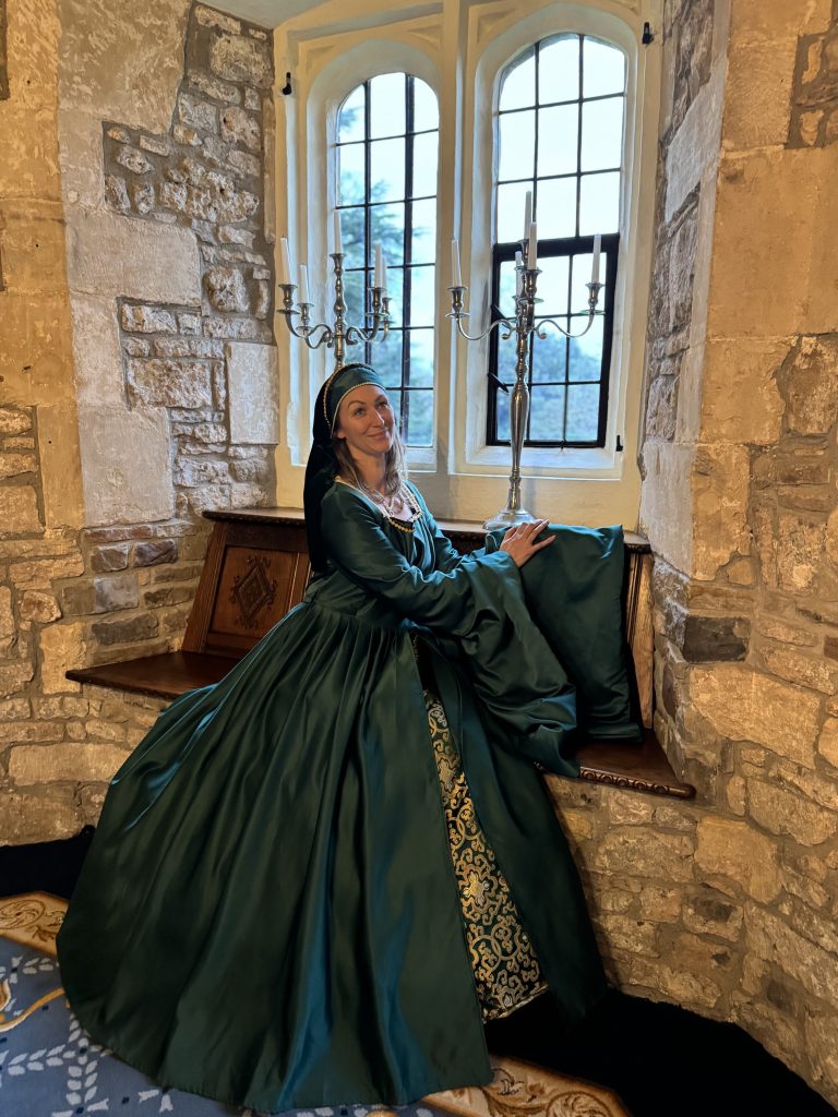 A person in a long green dress sits on a window seat in a stone room at Thornbury Castle, near a candelabra and arched windows, savoring the serenity of night in the castle.