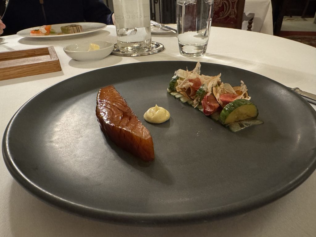 On a white tablecloth sits a dark round dish, showcasing glazed salmon, a dollop of sauce, and a salad with grapefruit and sliced vegetables—reminiscent of the regal feasts one might enjoy while spending a night at Thornbury Castle.
