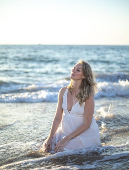 A woman in a white dress kneels on a beach at the water's edge with gentle waves in the background.