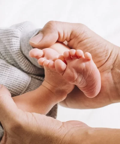 Adult hands gently holding a baby's feet, with the baby wearing gray knitted pants, and a white background.