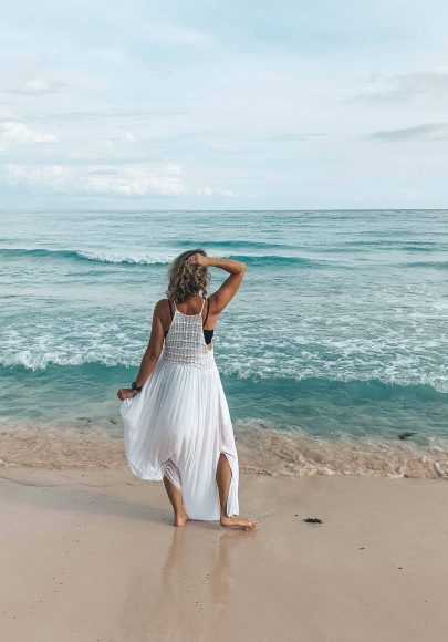 A person in a white dress stands on a sandy beach facing the ocean, with one hand on their head and waves gently crashing in the background.