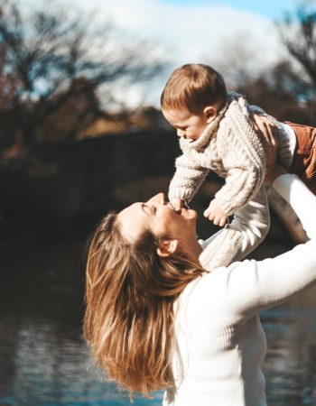 A woman in a white sweater lifts and plays with a baby near a body of water with autumn trees in the background.