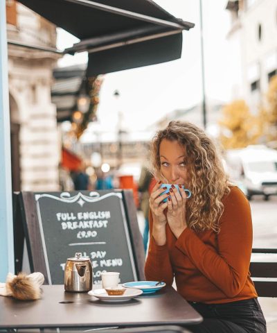 A woman with curly hair sits at an outdoor cafe table, sipping from a mug. There is a menu board behind her displaying breakfast options. Vehicles and buildings are visible in the background.