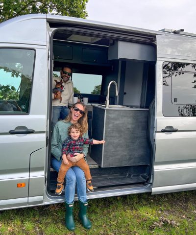 A family sits in and around a silver camper van parked on grass, with a man holding a small dog inside, a woman sitting on the step, and a child standing next to her.