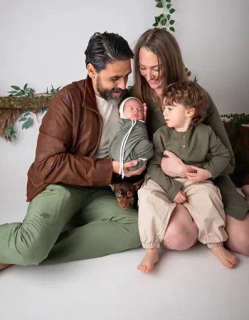 A seated family of four with a toddler and a newborn baby, along with a small dog under the father's arm, posing together in a studio filled with green and natural elements, gives a glimpse into their cozy "About Me" moment.