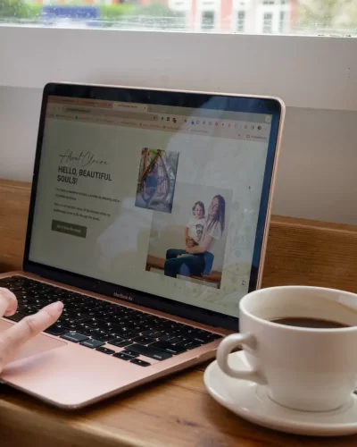 Person typing on a laptop displaying an "About Me" page on a wellness website beside a cup of coffee on a wooden table. Flowers in a vase are in the background.
