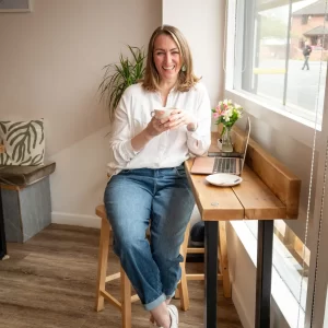 A woman in casual attire sits on a tall stool by a window, holding a coffee cup and smiling, with a laptop displaying her "About Me" page and a small vase of flowers on the wooden counter in front of her.