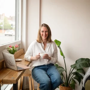 A smiling woman in a white shirt and jeans sits on a stool near a wooden desk with a laptop, holding a cup. There are plants and a window in the background, creating the perfect setting for an "About Me" introduction.