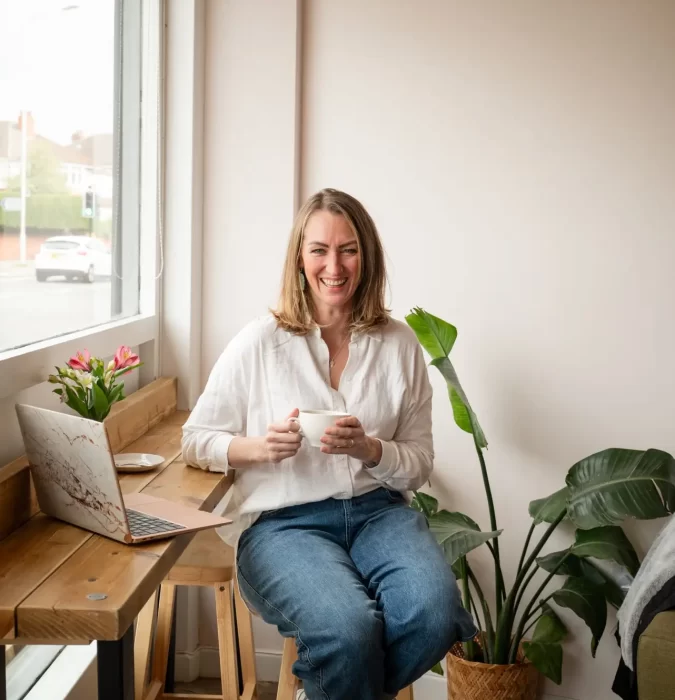A smiling woman in a white shirt and jeans sits on a stool near a wooden desk with a laptop, holding a cup. There are plants and a window in the background, creating the perfect setting for an "About Me" introduction.
