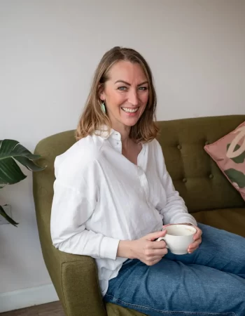 A woman with shoulder-length hair, wearing a white shirt and blue jeans, sits on a green sofa holding a white mug. In the background, there's a plant and a pillow that adds to the cozy "About Me" ambiance of her space.