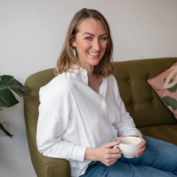 A woman with shoulder-length hair, wearing a white shirt and blue jeans, sits on a green sofa holding a white mug. In the background, there's a plant and a pillow that adds to the cozy "About Me" ambiance of her space.