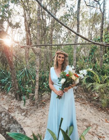 A woman in a light blue dress stands in a forest clearing, holding a bouquet of flowers, with the sun shining through the trees in the background.